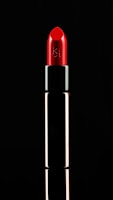 A vibrant red lipstick is displayed against a black background. The tube is metallic with a reflective surface. The lipstick is slightly angled, emphasizing its smooth and polished texture, with a logo etched on the side.