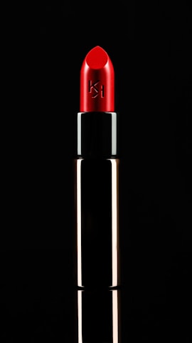 A vibrant red lipstick is displayed against a black background. The tube is metallic with a reflective surface. The lipstick is slightly angled, emphasizing its smooth and polished texture, with a logo etched on the side.