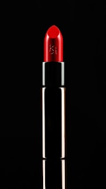 A vibrant red lipstick is displayed against a black background. The tube is metallic with a reflective surface. The lipstick is slightly angled, emphasizing its smooth and polished texture, with a logo etched on the side.
