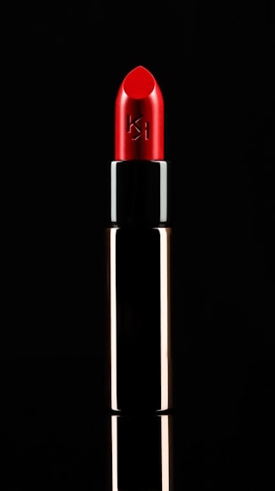 A vibrant red lipstick is displayed against a black background. The tube is metallic with a reflective surface. The lipstick is slightly angled, emphasizing its smooth and polished texture, with a logo etched on the side.