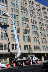 A tall beige building with multiple windows occupies the background. In the foreground, a large truck with an extendable platform is parked on the street. The platform is extended upward, reaching several stories high. Orange traffic cones are set up around the vehicle, and there is a person near the truck wearing a safety vest.