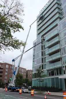 A glass-covered building is being cleaned or maintained using hydraulic boom lifts. Two platforms with workers are visible on the side of a high-rise structure. The scene is set in an urban environment with additional buildings visible in the background. Traffic cones and a small car are on the street below, suggesting maintenance work or caution.