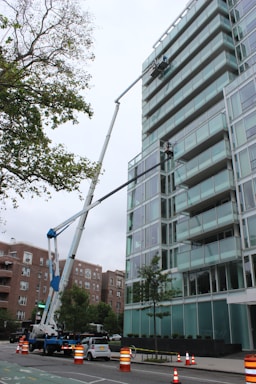 A glass-covered building is being cleaned or maintained using hydraulic boom lifts. Two platforms with workers are visible on the side of a high-rise structure. The scene is set in an urban environment with additional buildings visible in the background. Traffic cones and a small car are on the street below, suggesting maintenance work or caution.