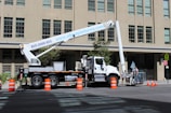 A large boom lift truck is parked on a city street next to a tall building. The truck has its arm extended with a platform at the end. Several orange traffic cones surround the vehicle, indicating an area of work or caution. Two workers appear to be standing on the platform of the boom lift, engaged in some sort of maintenance or construction activity.
