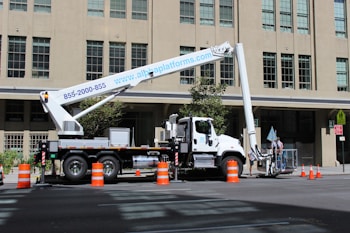 A large boom lift truck is parked on a city street next to a tall building. The truck has its arm extended with a platform at the end. Several orange traffic cones surround the vehicle, indicating an area of work or caution. Two workers appear to be standing on the platform of the boom lift, engaged in some sort of maintenance or construction activity.
