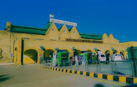 A historic railway station building with arched windows and a sign reading 'Rawalpindi Railway Station'. Several vehicles, including rickshaws and cars, are parked in front, with people walking around. The architecture is prominent with its yellow brick structure and green roof.