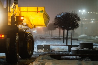 A large yellow snowplow is clearing snow from a dimly lit road. Decorative trees line the sidewalk, and string lights are visible in the background, creating a serene nighttime atmosphere. The area seems to be a public space, possibly a park or a pedestrian street.