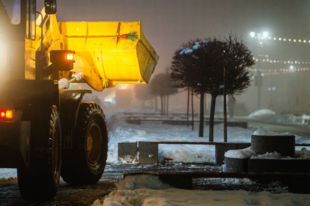 A large yellow snowplow is clearing snow from a dimly lit road. Decorative trees line the sidewalk, and string lights are visible in the background, creating a serene nighttime atmosphere. The area seems to be a public space, possibly a park or a pedestrian street.