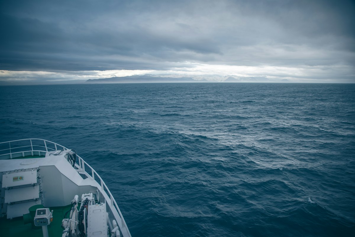 The white bow rail of a ship in one corner of the frame, dark blue open ocean ahead of it, heavy cloud, and a snow-covered strip of land on the horizon