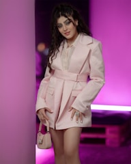 A person stands against a bright pink background, wearing a stylish light pink blazer with a matching belt and holding a small pale pink handbag. The outfit and background suggest a fashion-forward setting.