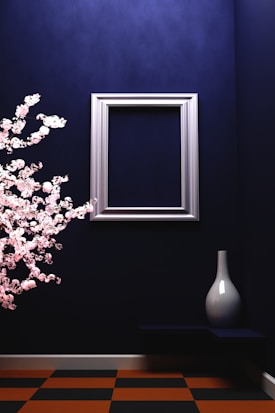 In a minimalist and modern setting, a silver picture frame is mounted on a deep blue wall. On the left, cherry blossom branches in full bloom add a touch of nature. On the right, a sleek white vase sits on a small shelf, contrasting against the dark background. The floor features a checkered pattern with alternating orange and black tiles.