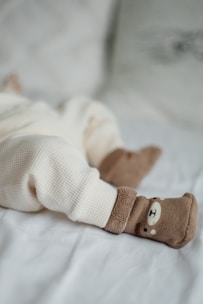 A cozy scene with a baby's legs dressed in soft cream-colored, textured pants lying on a light blanket. The feet are adorned with adorable brown socks featuring a cute bear face design.
