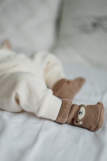 A cozy scene with a baby's legs dressed in soft cream-colored, textured pants lying on a light blanket. The feet are adorned with adorable brown socks featuring a cute bear face design.