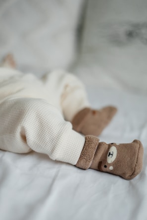 A cozy scene with a baby's legs dressed in soft cream-colored, textured pants lying on a light blanket. The feet are adorned with adorable brown socks featuring a cute bear face design.