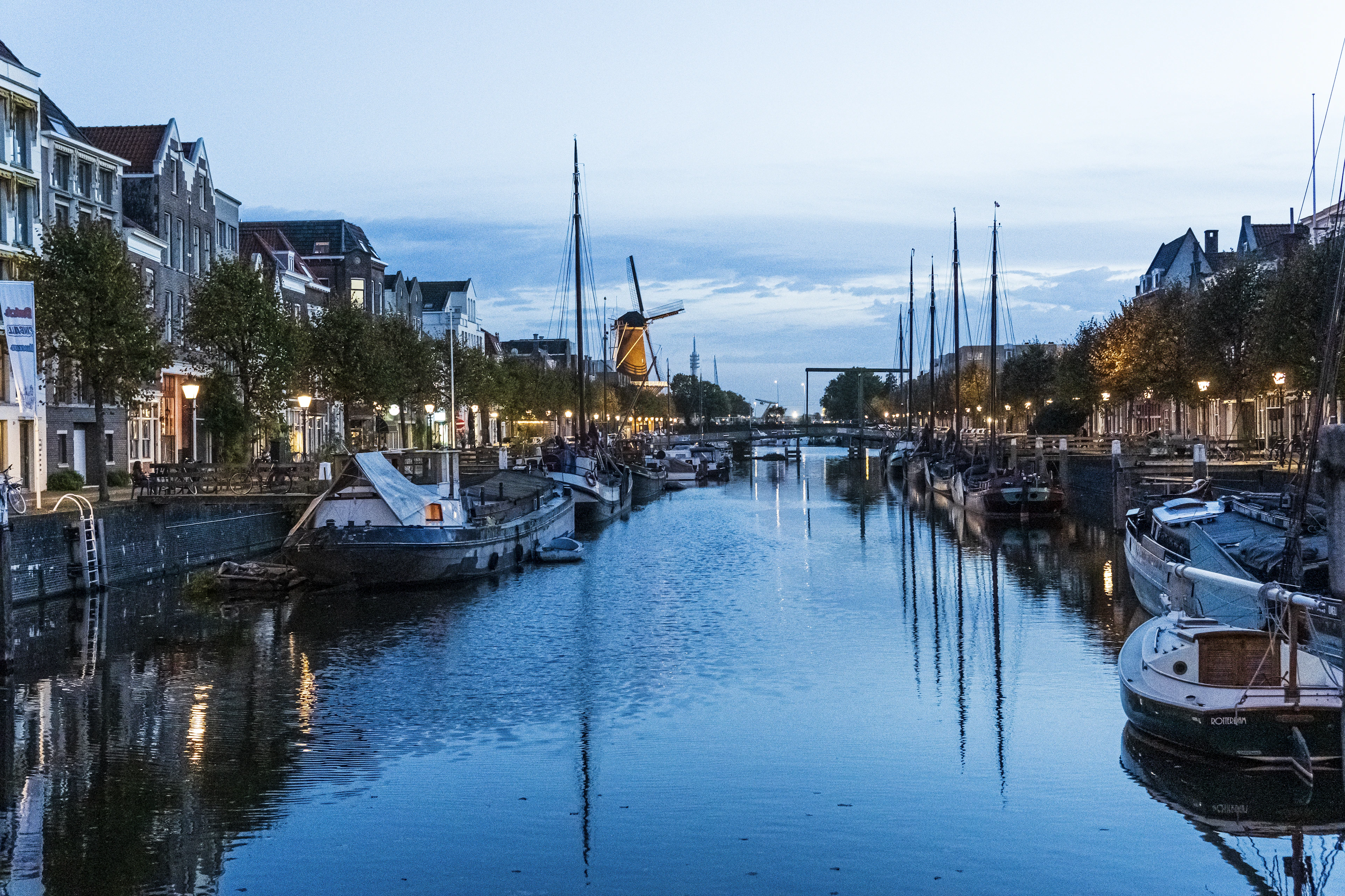 Historic masted barges moored along the Delfshaven canal in Rotterdam at blue hour, lit quayside windows and street lamps reflected in still water with a windmill silhouetted at the far end.