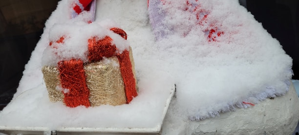 A snow-covered gift box wrapped in red and gold sits on a white surface. The gift is surrounded by fake snow and fluffy white fabric, accented with touches of red.