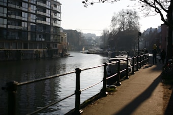 A tranquil riverside scene with a modern apartment building on the left, bordered by calm water and a pathway on the right. The path is flanked by a railing and lined with bare trees, casting long shadows in the soft light. Several people are strolling along the walkway, some with dogs, creating a peaceful urban atmosphere. Boats are moored in the distance under a hazy sky with hills visible.