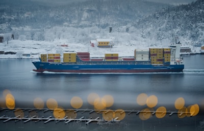 A large container ship with a blue hull travels through a calm, snow-covered landscape. Stacked colorful containers are visible on the ship as it navigates through a narrow body of water. The surrounding scenery features snowy hillsides with sparse trees and a few vibrant houses, creating a contrast against the white snow. In the foreground, there are blurred, warm-colored circular lights, adding depth to the image.