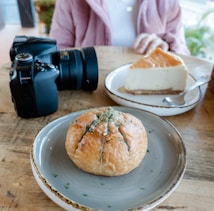 A wooden table with a camera, a round bread with herbs on a gray plate, and a slice of cheesecake on a white plate with a spoon. A person wearing a pink jacket sits in the background.