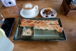 A wooden table holds a plate with two breadsticks garnished with herbs and accompanied by a creamy dipping sauce. Nearby, a cup of cappuccino features latte art resembling an animal face. A small dessert plate contains a slice of chocolate cake garnished with orange slices and drizzled with syrup. A smartphone is placed beside a glass of water with a lemon slice.