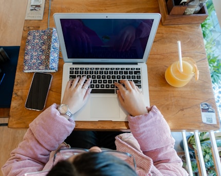 A person seated at a wooden table is working on a laptop, with hands on the keyboard. Beside the laptop, there is a floral notebook, a smartphone, and a glass of orange juice with a straw. The person is wearing a pink, textured jacket and a silver wristwatch.