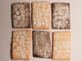 A set of six rectangular cookies arranged in two rows of three on a light background. The cookies have intricate floral patterns made of powdered sugar on their surfaces. There are two types, one with a darker brown color and another with a lighter golden-brown hue.