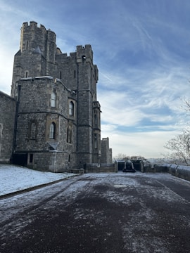 A large, historic stone castle with multiple tall, rectangular towers stands prominently. The exterior is made of grey stone bricks with arched windows. A lightly snow-covered pathway leads to a cemented platform where a cannon is positioned, facing outward. The sky is bright with wispy clouds, and bare trees are visible on the right side.