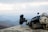 A tactical helmet with attached night vision equipment rests on a rocky surface against a backdrop of a hazy mountainous landscape. The helmet is equipped with various attachments, including a mounted camera and communication devices.