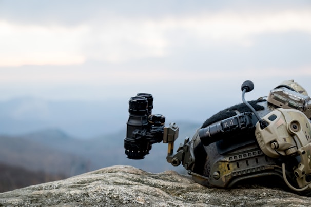 A tactical helmet with attached night vision equipment rests on a rocky surface against a backdrop of a hazy mountainous landscape. The helmet is equipped with various attachments, including a mounted camera and communication devices.