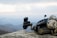 A tactical helmet with attached night vision equipment rests on a rocky surface against a backdrop of a hazy mountainous landscape. The helmet is equipped with various attachments, including a mounted camera and communication devices.