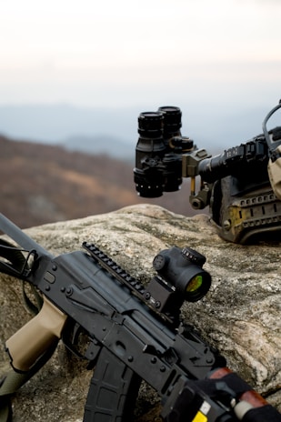 A rifle is laid on a rocky surface with a sight attachment visible. Nearby, a set of binoculars and a tactical helmet are placed. The background shows a blurry, muted landscape with a distant horizon.