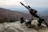 A rocky terrain serves as the foreground, featuring tactical military gear including a helmet with a camouflage pattern, night vision goggles, and a rifle equipped with sights and other attachments. The background reveals a misty, mountainous landscape under an overcast sky.