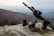 A rocky terrain serves as the foreground, featuring tactical military gear including a helmet with a camouflage pattern, night vision goggles, and a rifle equipped with sights and other attachments. The background reveals a misty, mountainous landscape under an overcast sky.