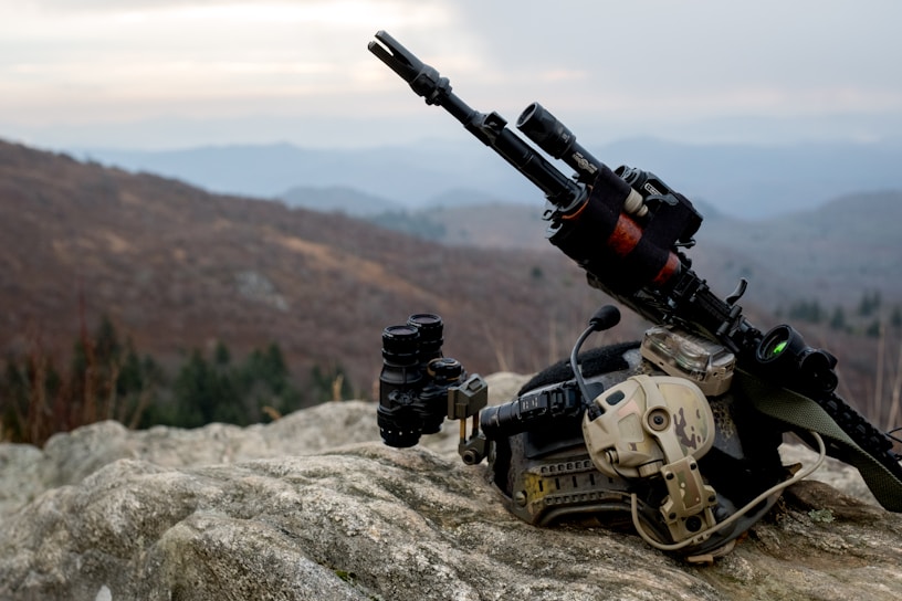 A rocky terrain serves as the foreground, featuring tactical military gear including a helmet with a camouflage pattern, night vision goggles, and a rifle equipped with sights and other attachments. The background reveals a misty, mountainous landscape under an overcast sky.