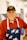 An elderly man is smiling warmly, wearing a hat with the text 'Vietnam Veteran' and a red zip-up jacket. He has a white apron with a pattern of stars. The background seems to be indoors, slightly blurred.