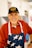 An elderly man is smiling warmly, wearing a hat with the text 'Vietnam Veteran' and a red zip-up jacket. He has a white apron with a pattern of stars. The background seems to be indoors, slightly blurred.