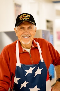 An elderly man is smiling warmly, wearing a hat with the text 'Vietnam Veteran' and a red zip-up jacket. He has a white apron with a pattern of stars. The background seems to be indoors, slightly blurred.