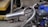 A close-up view of a small engine with a curved metal exhaust and a tire in the foreground. The engine appears to be mounted on a framework with a chain drive system. In the background, there are metal structures and a yellow trolley.
