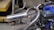 A close-up view of a small engine with a curved metal exhaust and a tire in the foreground. The engine appears to be mounted on a framework with a chain drive system. In the background, there are metal structures and a yellow trolley.