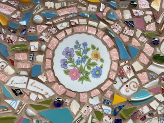 A mosaic-style artwork featuring a central decorative plate with hand-painted flowers in pink, blue, and yellow. Surrounding the plate are numerous small, colorful ceramic and tile pieces, including shades of pink, blue, green, and yellow, creating an intricate pattern. Some pieces have floral designs, while others are solid colors or have abstract patterns.