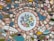 A mosaic-style artwork featuring a central decorative plate with hand-painted flowers in pink, blue, and yellow. Surrounding the plate are numerous small, colorful ceramic and tile pieces, including shades of pink, blue, green, and yellow, creating an intricate pattern. Some pieces have floral designs, while others are solid colors or have abstract patterns.