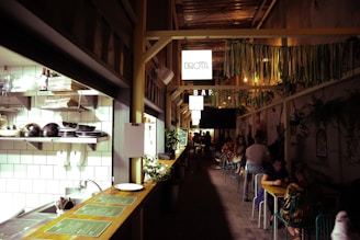 A narrow, stylish cafe corridor with an open kitchen section on the left. Shelves are stocked with plates and bowls, and there's greenery alongside the counter. On the right, people are seated at small tables along the wall, which is adorned with plants and decorative elements. Overhead, there are hanging signs and colorful ribbons adding to the cozy and vibrant atmosphere.