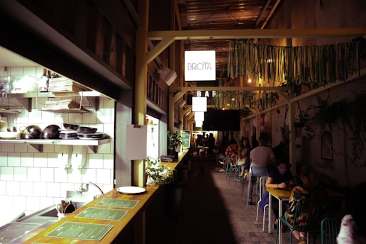 A narrow, stylish cafe corridor with an open kitchen section on the left. Shelves are stocked with plates and bowls, and there's greenery alongside the counter. On the right, people are seated at small tables along the wall, which is adorned with plants and decorative elements. Overhead, there are hanging signs and colorful ribbons adding to the cozy and vibrant atmosphere.