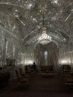 An opulent room with intricately designed mirrored walls and ceiling. The central chandelier casts a warm glow, enhancing the elaborate patterns. Luxurious drapery covers a large window, and a collection of ornate chairs and a sofa occupy the room. A dark bust is visible on a side table, contributing to the elegant atmosphere.