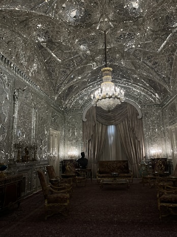 An opulent room with intricately designed mirrored walls and ceiling. The central chandelier casts a warm glow, enhancing the elaborate patterns. Luxurious drapery covers a large window, and a collection of ornate chairs and a sofa occupy the room. A dark bust is visible on a side table, contributing to the elegant atmosphere.