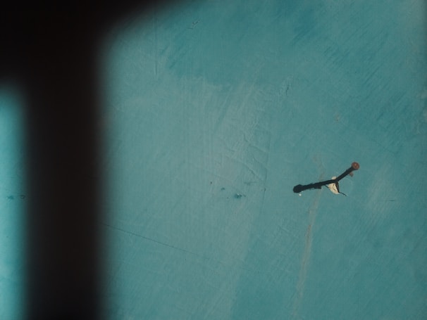 A single rusty nail protrudes from a teal-colored surface, casting a long shadow across the textured background. A hint of paper or a similar material is caught on the nail, adding to the scene.
