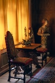 A richly decorated interior features two ornate chairs with red and gold upholstery, placed beside a wooden table. A model ship decorates the tabletop, illuminated by soft, warm light streaming through mustard yellow curtains. Nearby, a classical sculpture bust rests on a pedestal, surrounded by intricate wooden paneling and a patterned carpet.