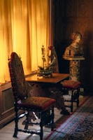 A richly decorated interior features two ornate chairs with red and gold upholstery, placed beside a wooden table. A model ship decorates the tabletop, illuminated by soft, warm light streaming through mustard yellow curtains. Nearby, a classical sculpture bust rests on a pedestal, surrounded by intricate wooden paneling and a patterned carpet.