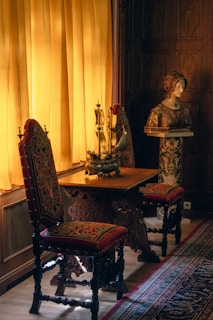 A richly decorated interior features two ornate chairs with red and gold upholstery, placed beside a wooden table. A model ship decorates the tabletop, illuminated by soft, warm light streaming through mustard yellow curtains. Nearby, a classical sculpture bust rests on a pedestal, surrounded by intricate wooden paneling and a patterned carpet.