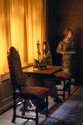 A richly decorated interior features two ornate chairs with red and gold upholstery, placed beside a wooden table. A model ship decorates the tabletop, illuminated by soft, warm light streaming through mustard yellow curtains. Nearby, a classical sculpture bust rests on a pedestal, surrounded by intricate wooden paneling and a patterned carpet.